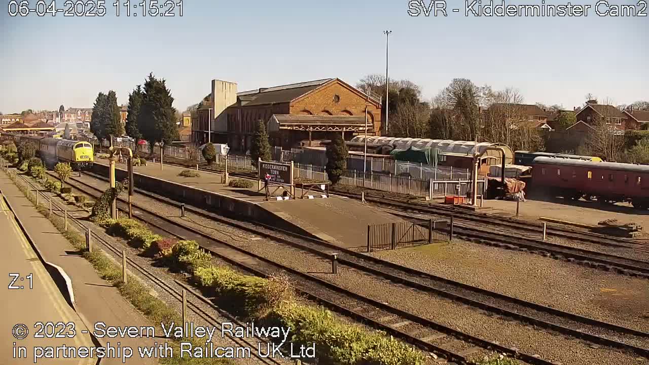 A sunny day at a train station shows a yellow and gray train departing from a platform, with several other railcars and a brick station building in the background.