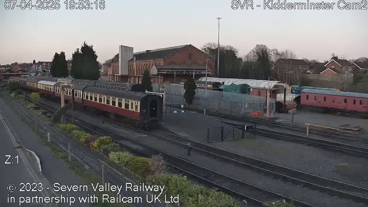 A train of vintage passenger cars sits at a station on railway tracks under a twilight sky.