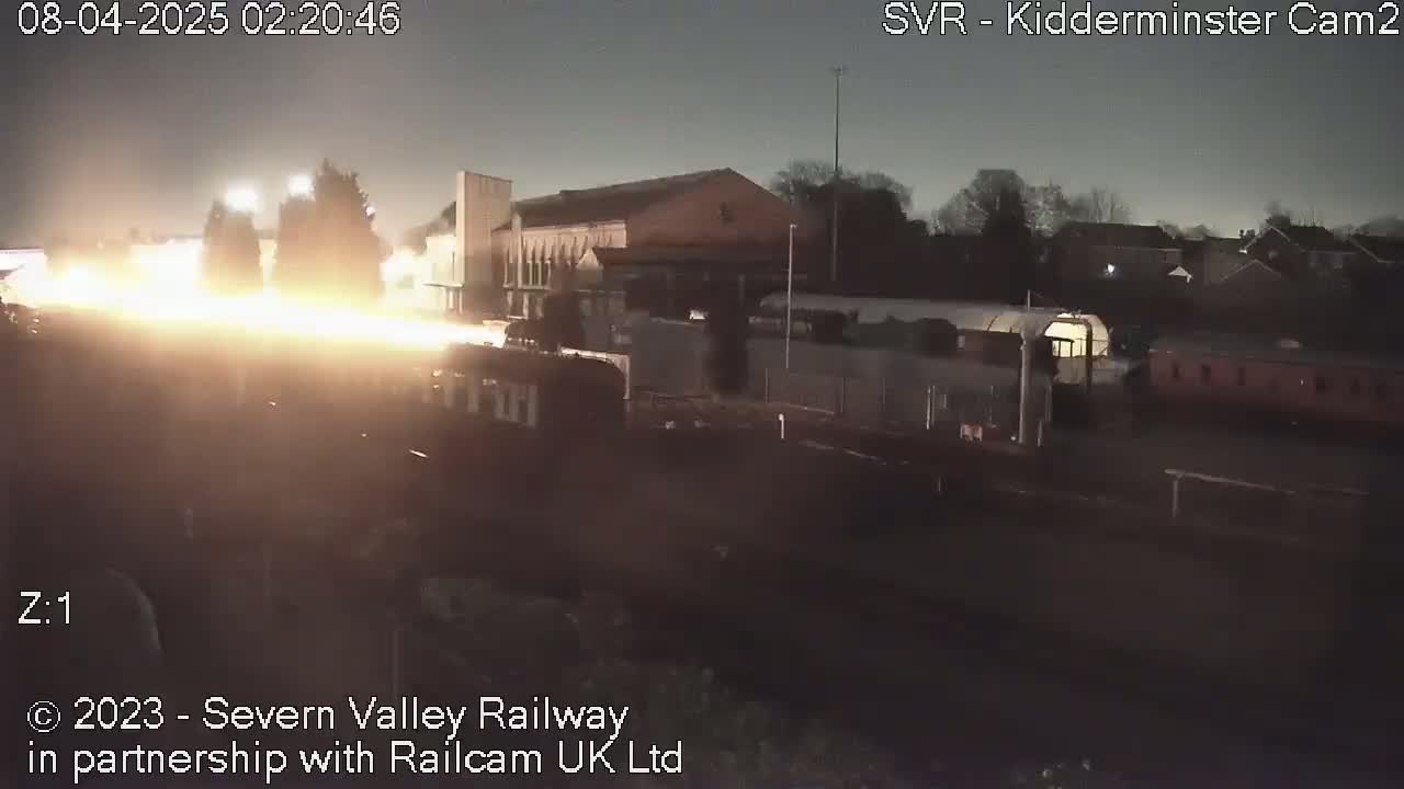A nighttime view of a train moving past buildings under a dark sky.