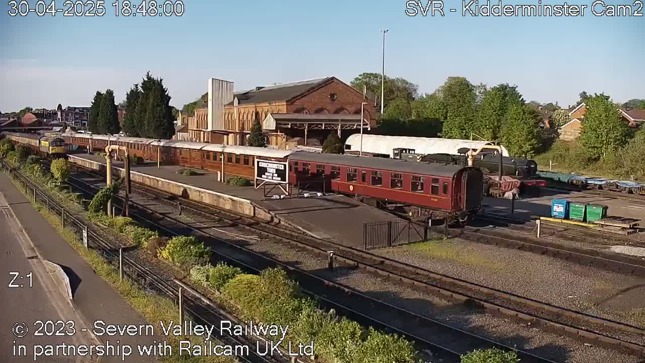 A sunny day at a train station shows several passenger cars and locomotives on the tracks, some parked and others appearing to be in motion.