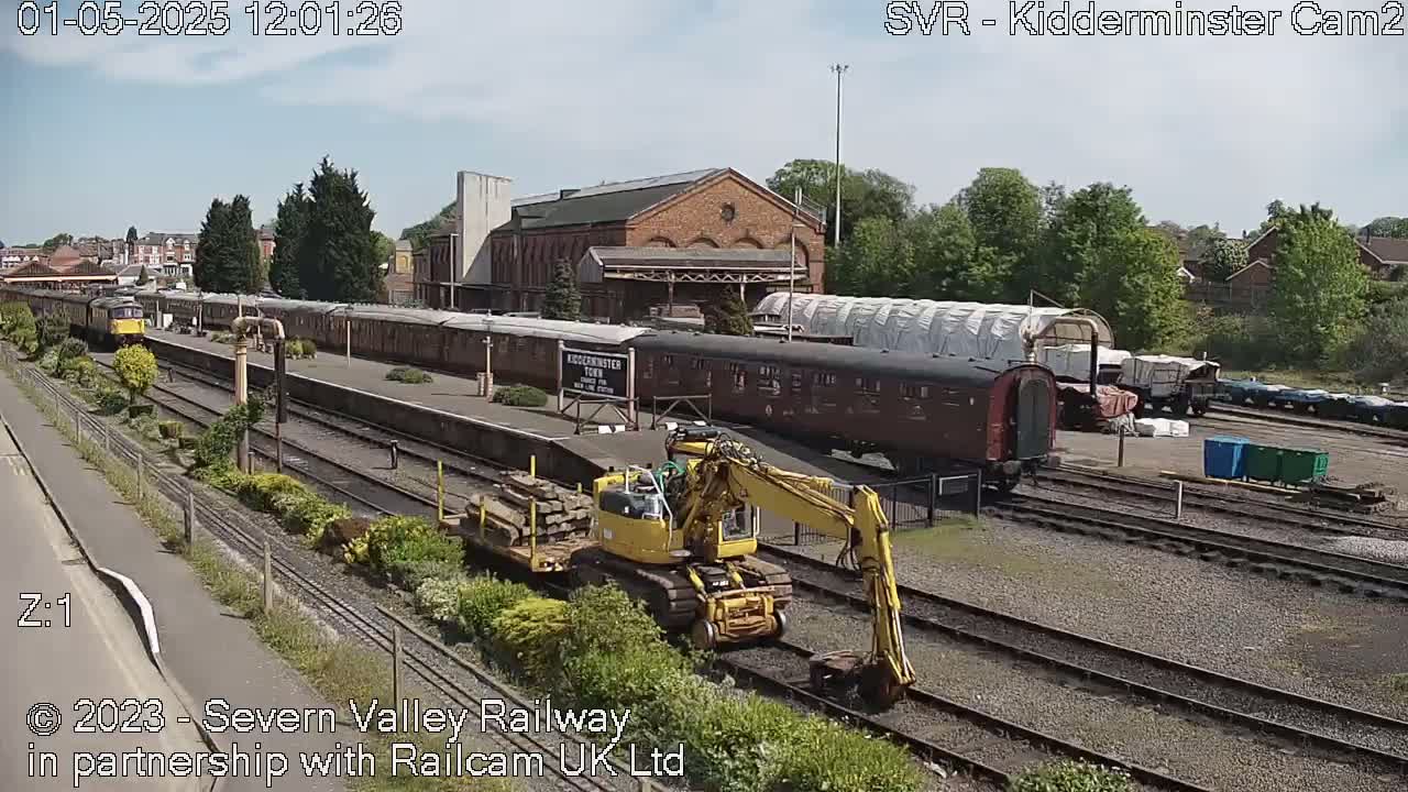 A yellow excavator sits on train tracks next to a train station on a sunny day, with multiple train cars and a locomotive visible.