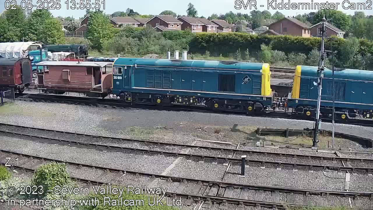 A teal and yellow diesel locomotive and several railway cars sit on train tracks in front of a residential area on a sunny day.