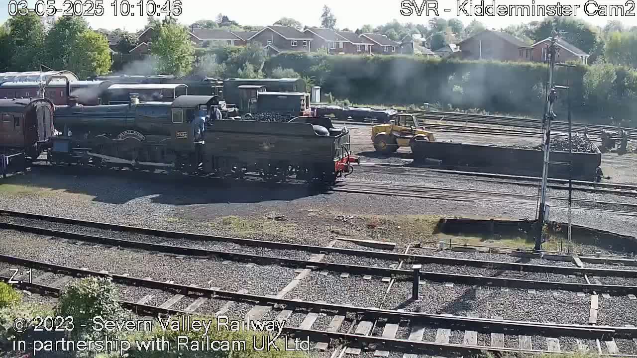 A steam locomotive and several rail cars sit on a railway track in sunny weather, with a yellow digger visible nearby.