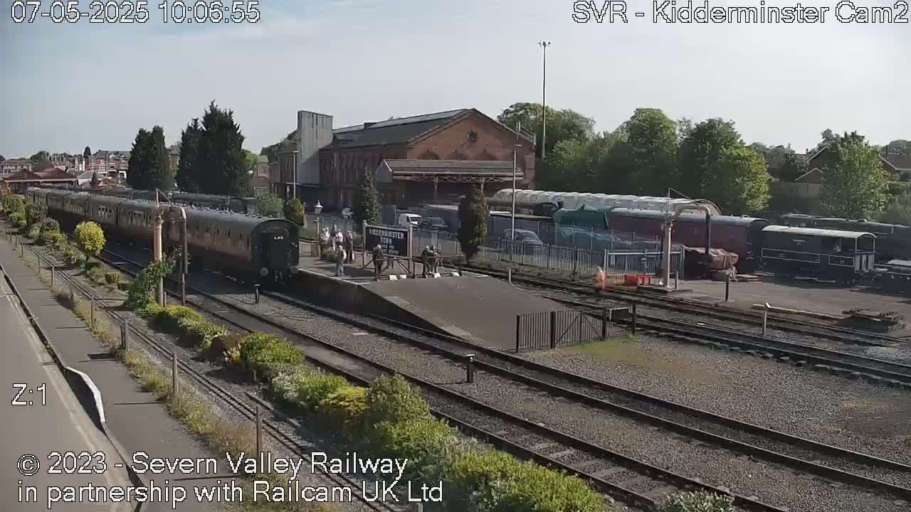 A long passenger train pulls into a station on a sunny day, with other train cars visible in the background.