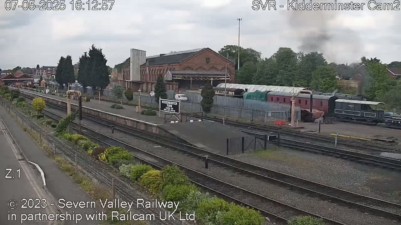 A train station with several railway cars and a brick building is visible under an overcast sky.