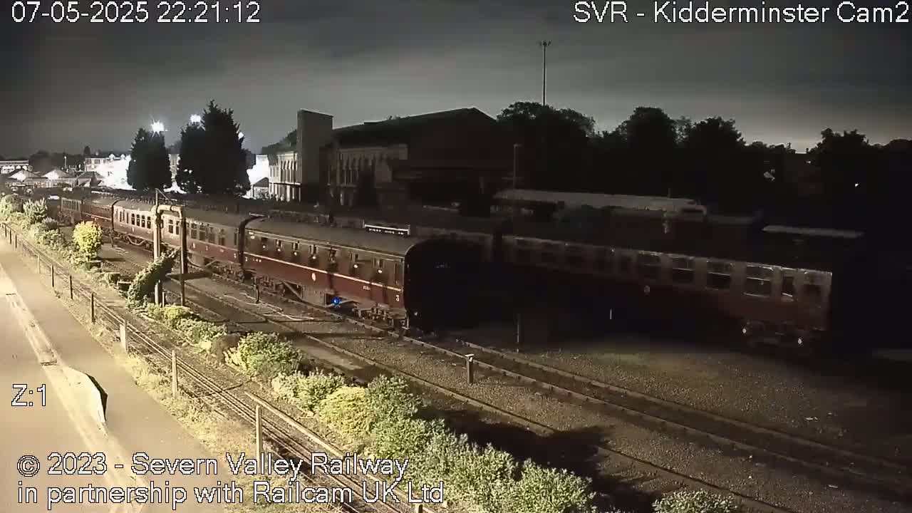 A long maroon passenger train sits stationary on railway tracks at night under a dark, overcast sky.