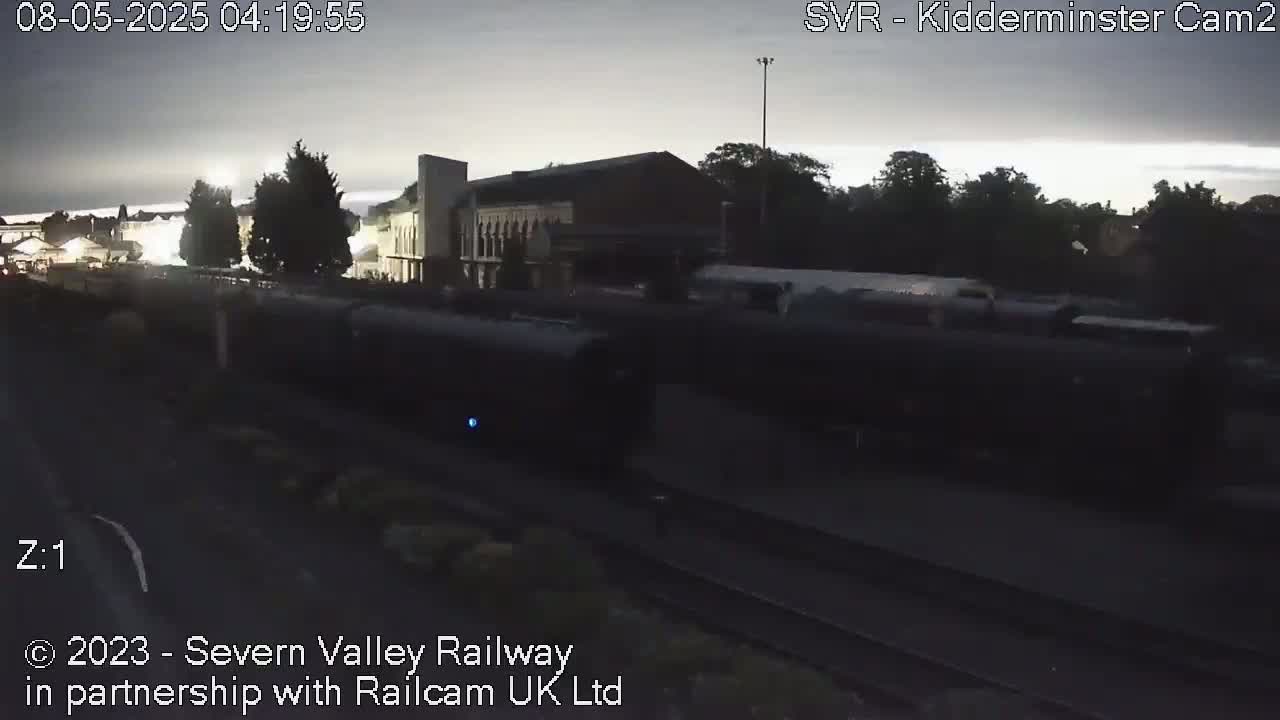 A long dark train is moving past a station under an overcast sky at dawn.