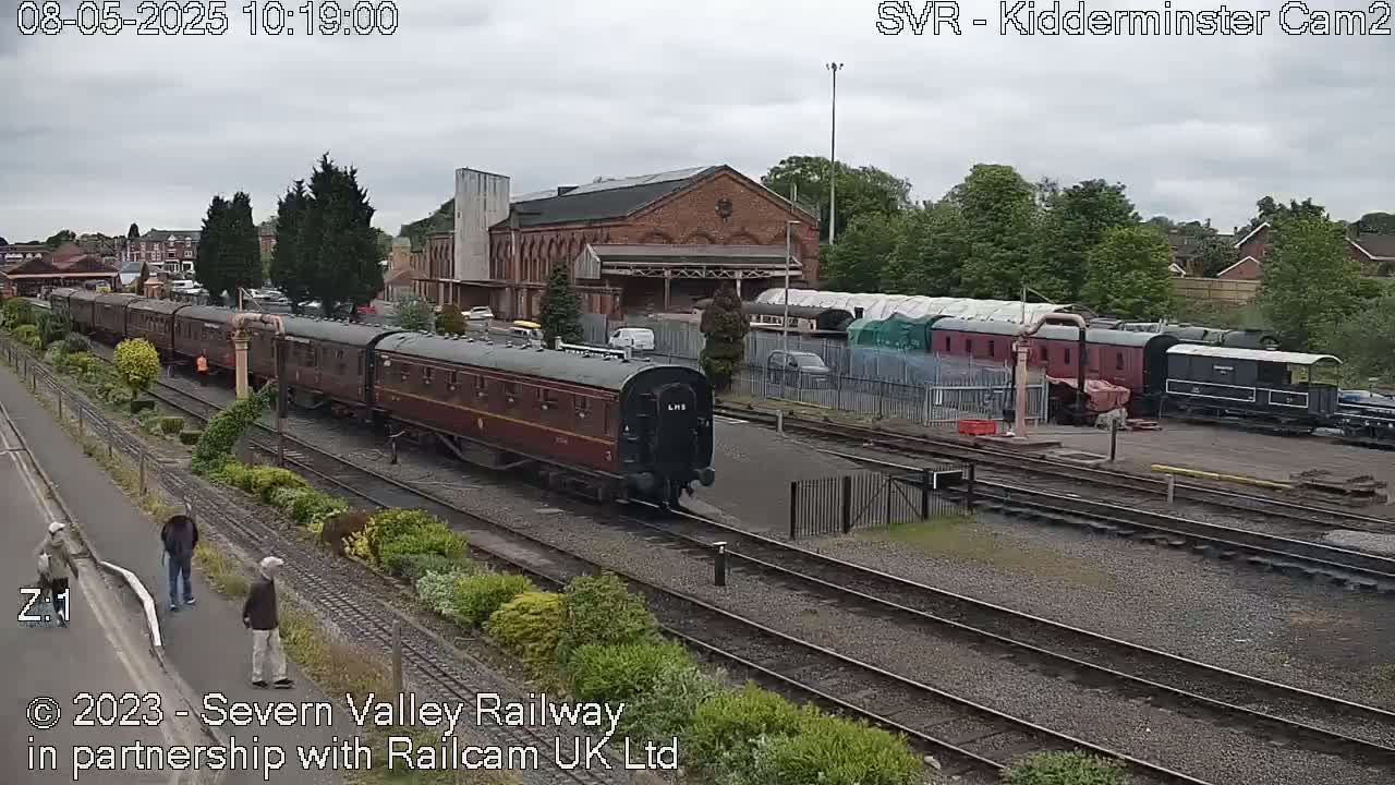 A long train of maroon carriages pulled by a black locomotive is moving along railway tracks under an overcast sky, past a railway station and other parked trains.