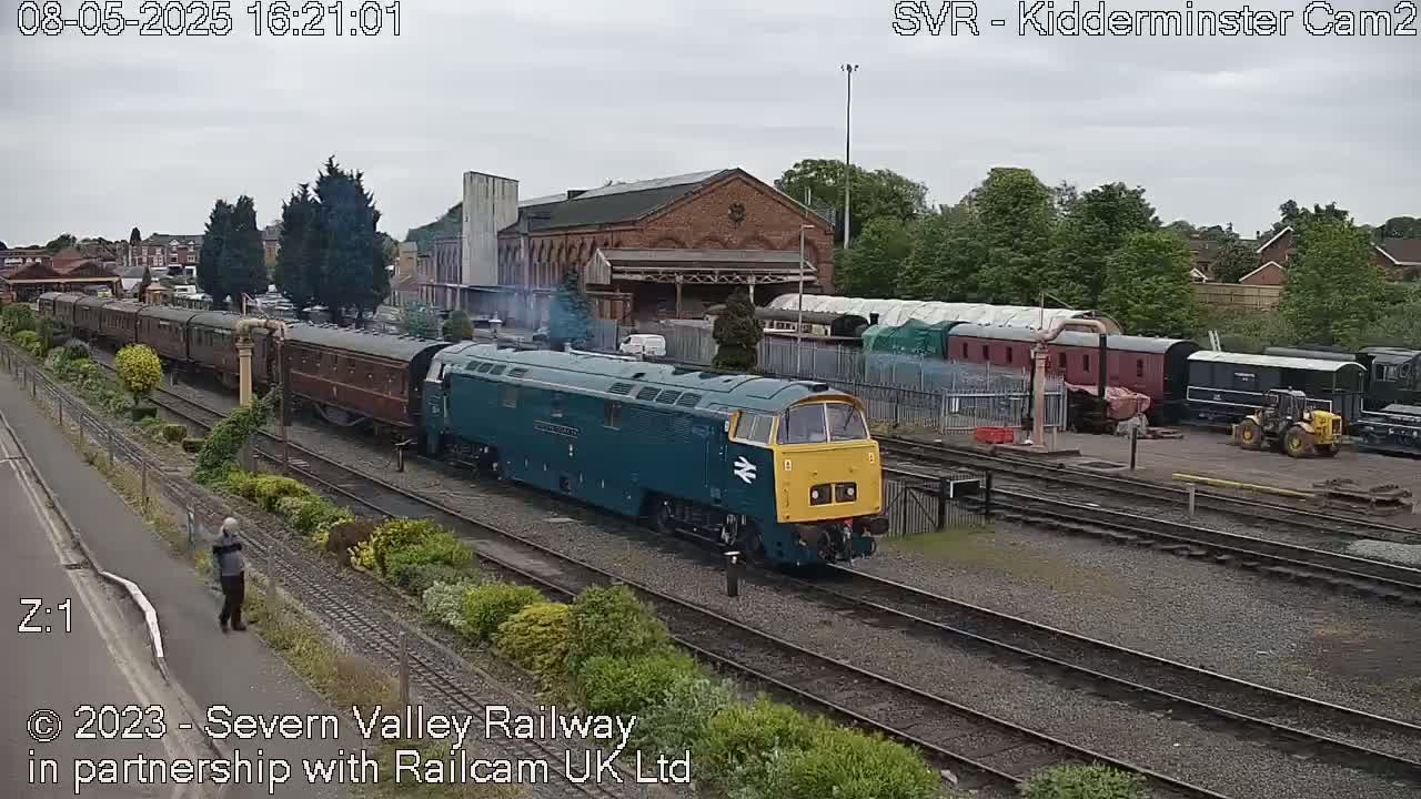 A teal and yellow diesel locomotive pulls a long train of passenger cars on a railway track under an overcast sky.