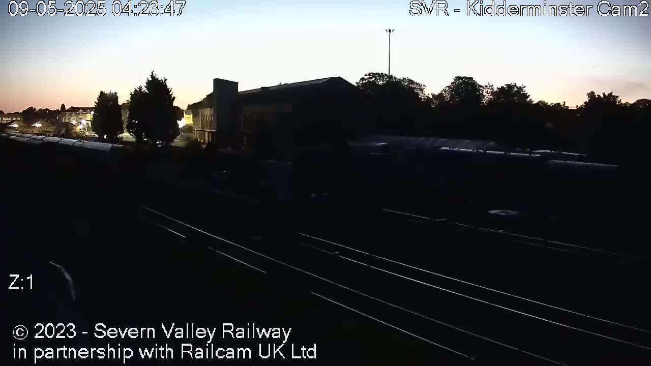 A nighttime, pre-dawn view of a train station with several railcars and a building silhouetted against a light sky.