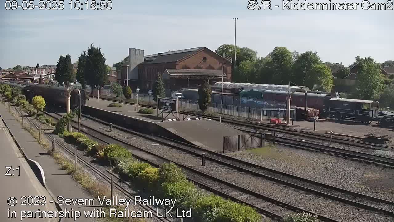 A sunny day at a train station shows multiple train cars and a locomotive on the tracks near a brick building.