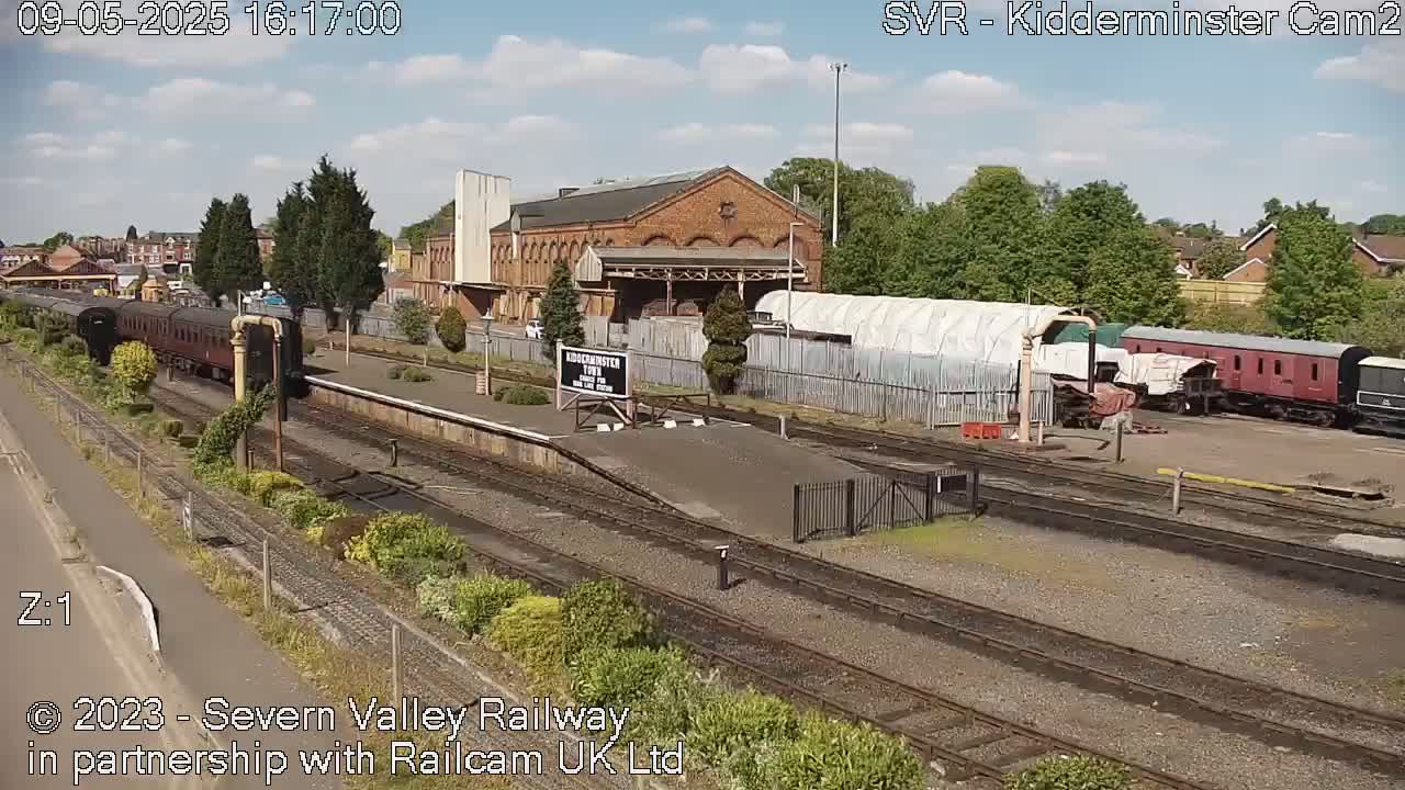 A partly cloudy day at a train station shows a long passenger train leaving a brick building, with several other train cars parked nearby.