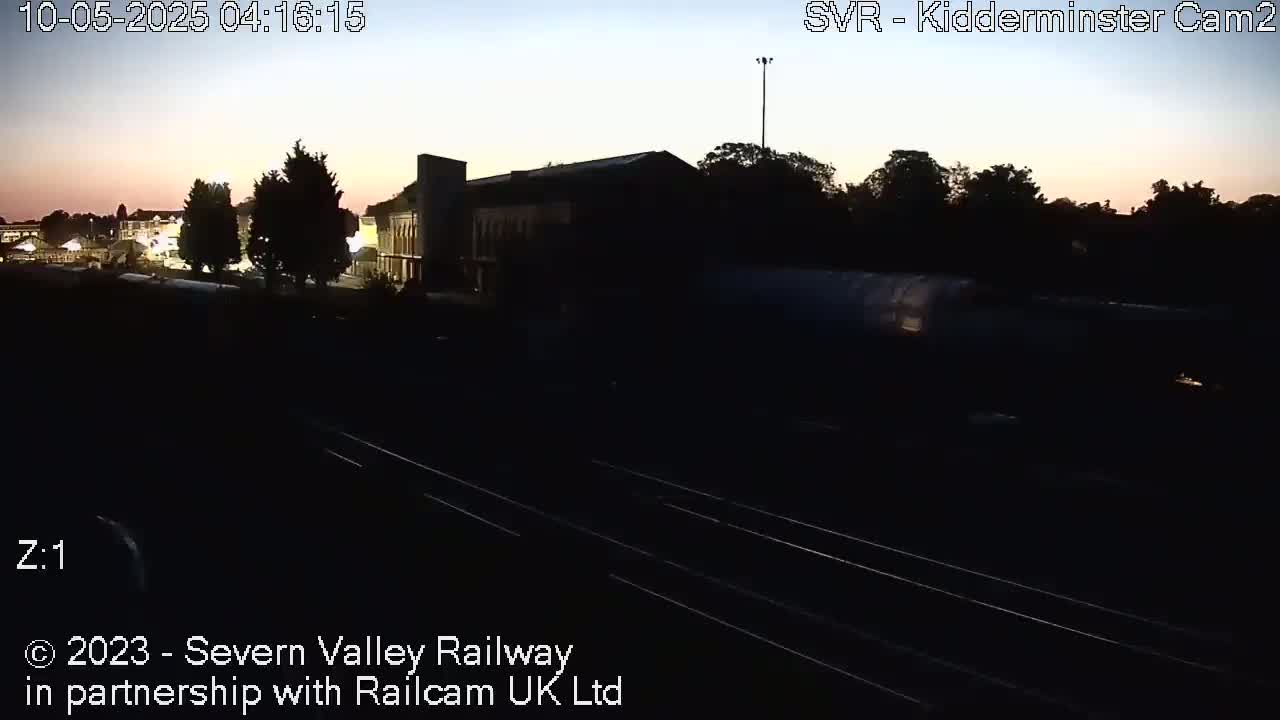 A dark, pre-dawn scene shows a train passing a railway station building and trees under a twilight sky.
