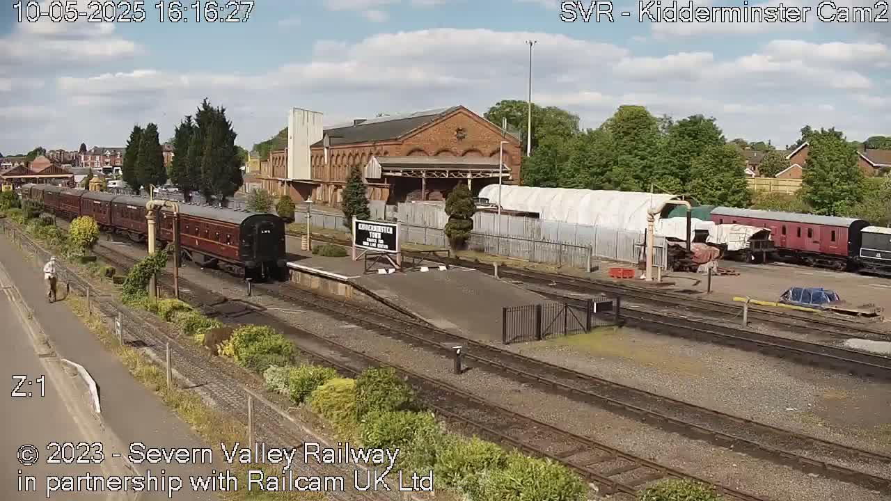 A long passenger train sits at a railway station on a partly sunny day, with other train cars parked nearby and a person walking on a path.