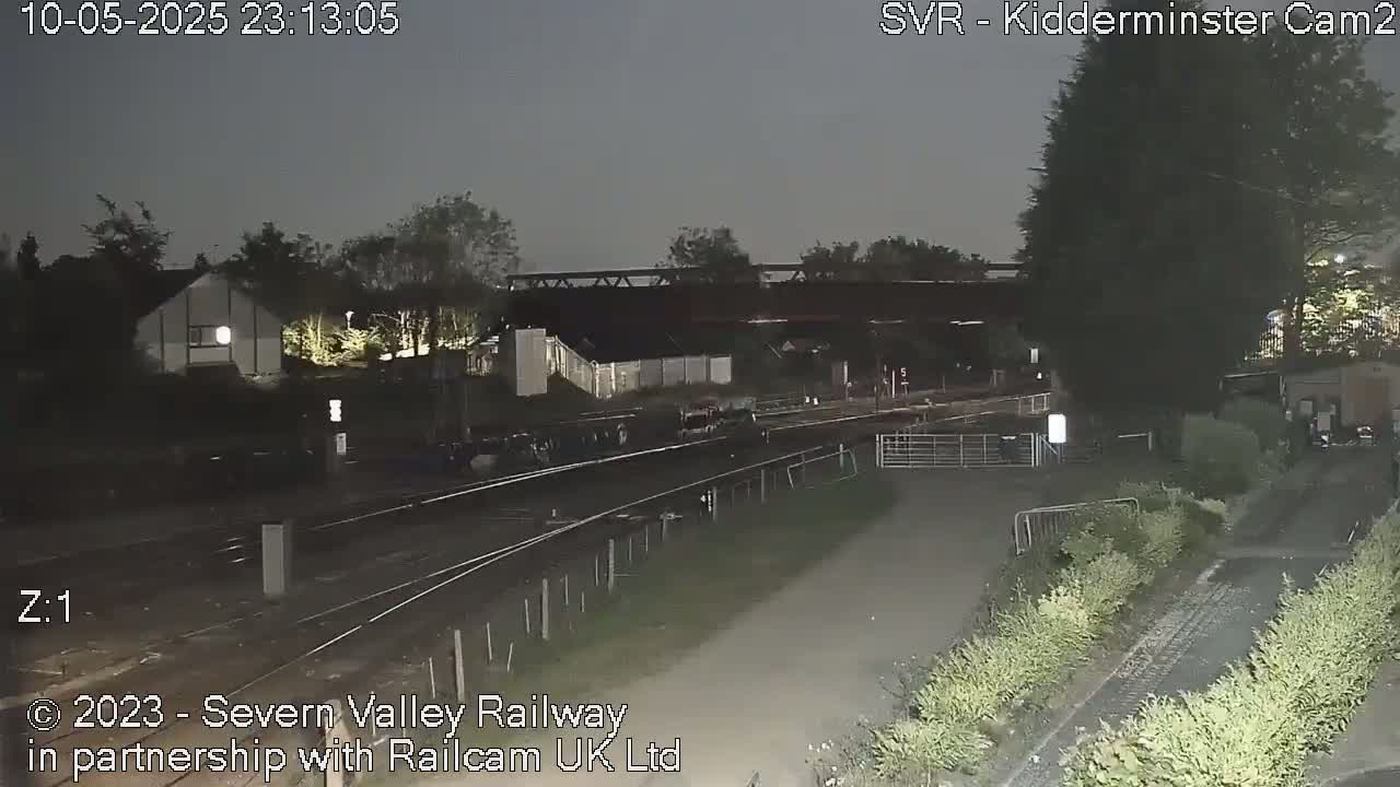 A nighttime view of railway tracks, a metal bridge, and surrounding buildings and vegetation under a dark sky.