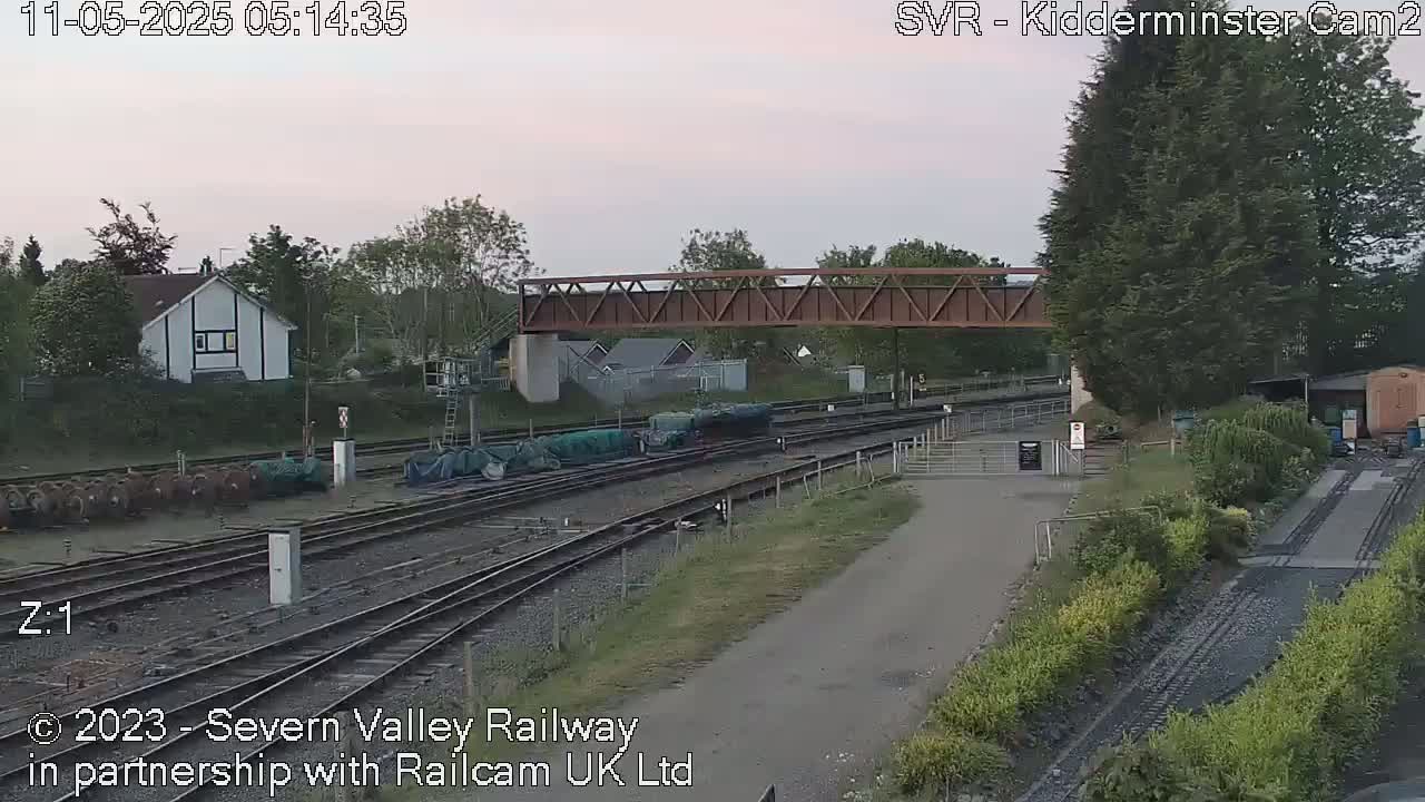Under an overcast sky, a railway yard with multiple tracks, a footbridge, and several railway cars is shown.