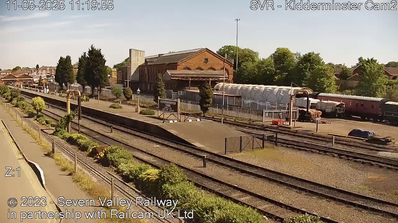 A sunny day at a train station with multiple tracks, a brick building, and several train cars parked nearby.