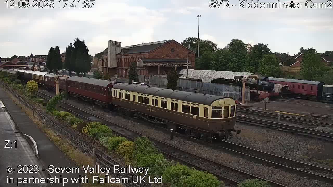 A long train of vintage passenger cars sits on railway tracks under an overcast sky, with additional rail cars and buildings visible in the background.