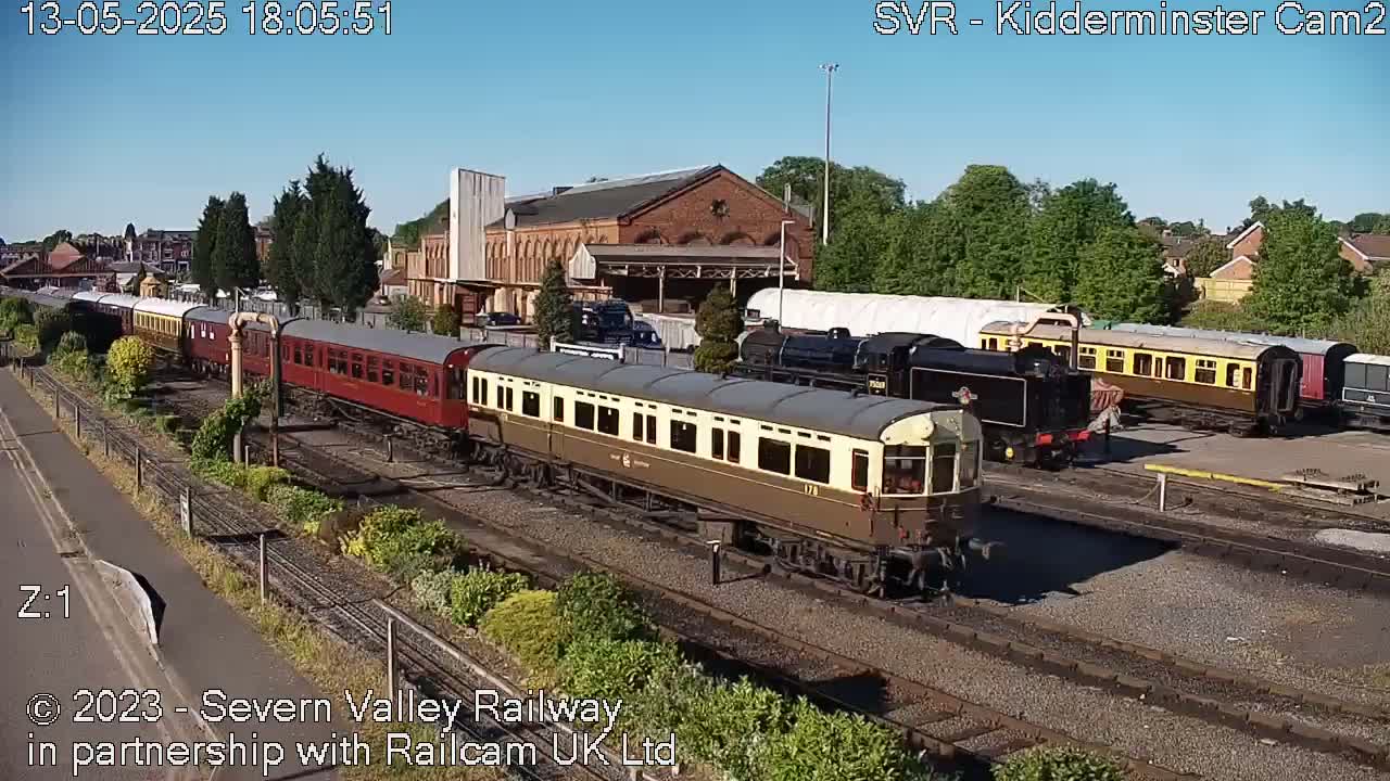 Under a clear blue sky, several vintage railway carriages and locomotives are parked on a railway track next to a brick building.