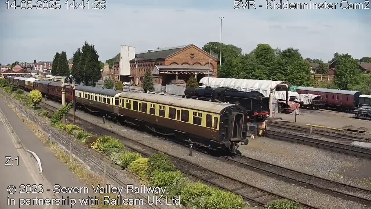 A long train of vintage passenger cars sits on railway tracks in front of a brick building under a partly sunny sky.