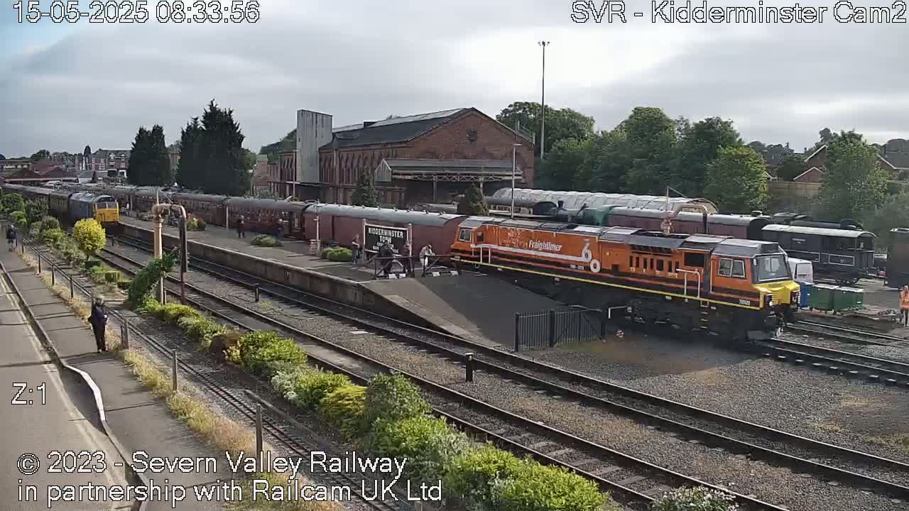 A long train of carriages and a diesel locomotive sits at a train station on an overcast day.