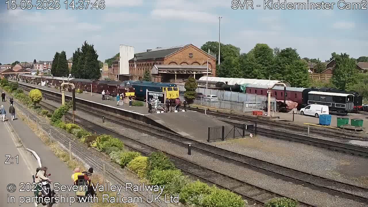 A sunny day at a train station shows multiple passenger train cars and a diesel locomotive, with people standing on the platform and others walking along a nearby path.