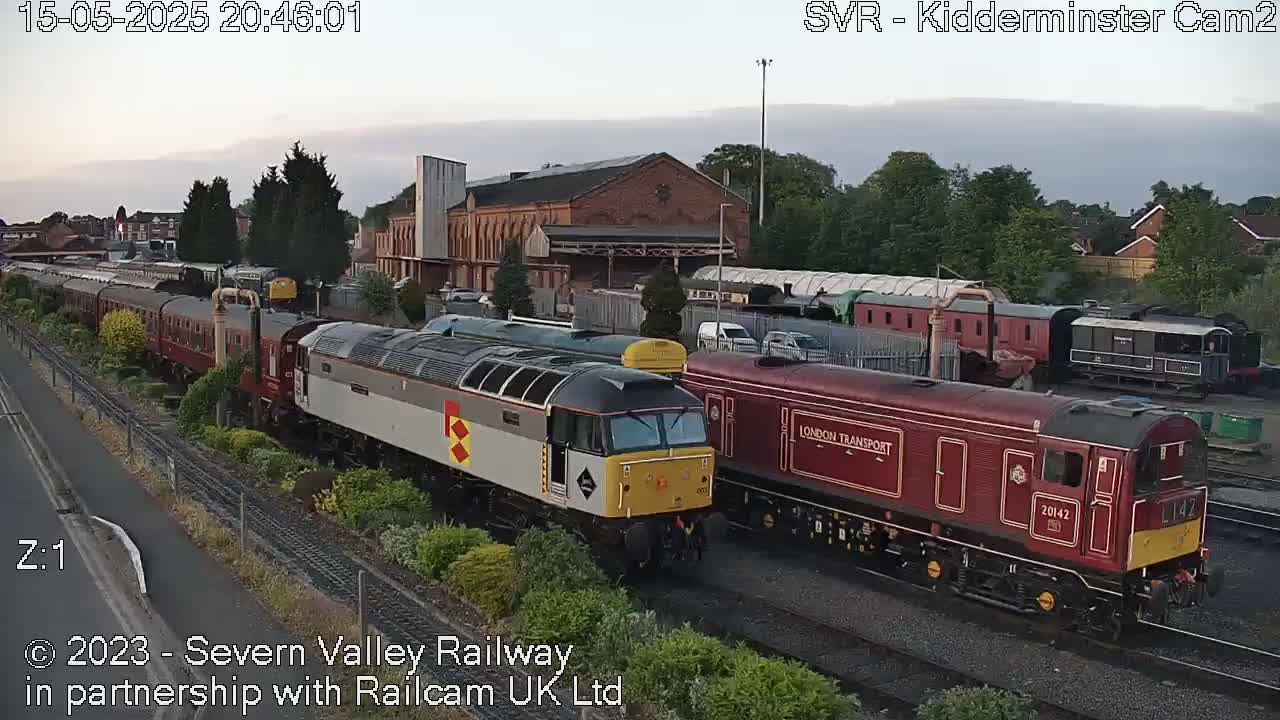 Under a twilight sky, several diesel locomotives and passenger cars are parked on railway tracks near a brick building.