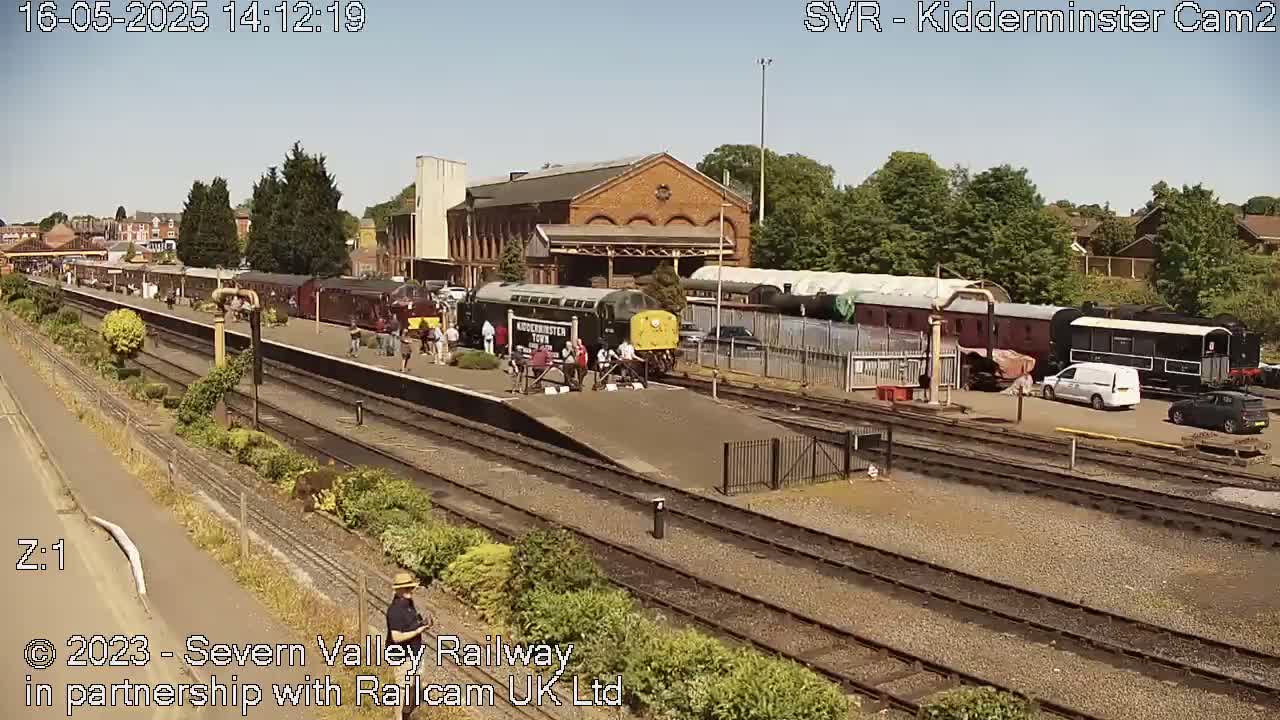 A sunny day at a train station shows multiple train cars and a locomotive parked on several tracks, with people standing around the train and cars parked nearby.