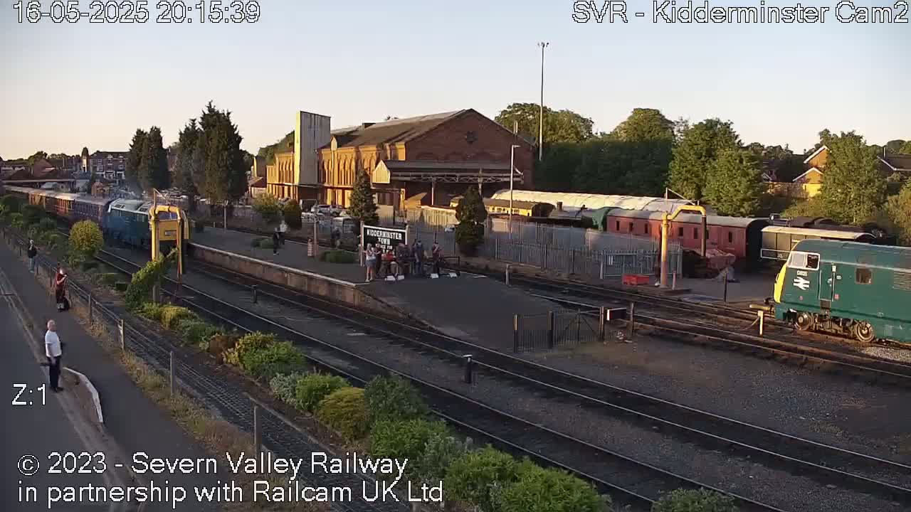 A passenger train pulls into a railway station at sunset, with several other train cars and people visible on the platform.