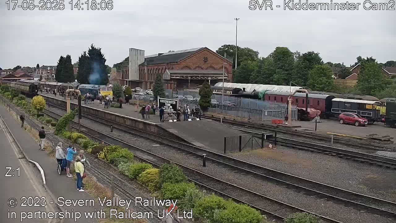 A steam train is arriving at a train station on an overcast day, with several people gathered on the platform and parked cars visible nearby.