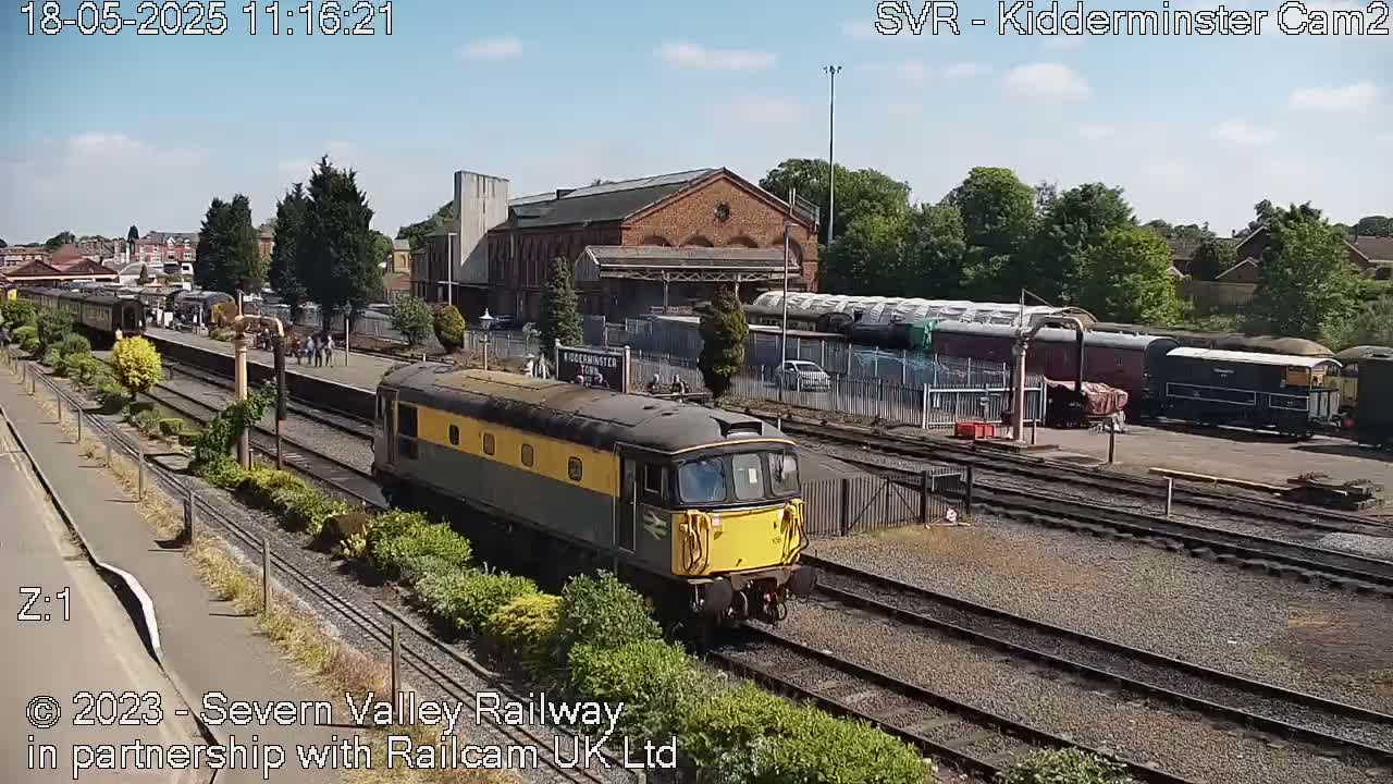 A green and yellow diesel locomotive leads a train through a railway station on a sunny day.