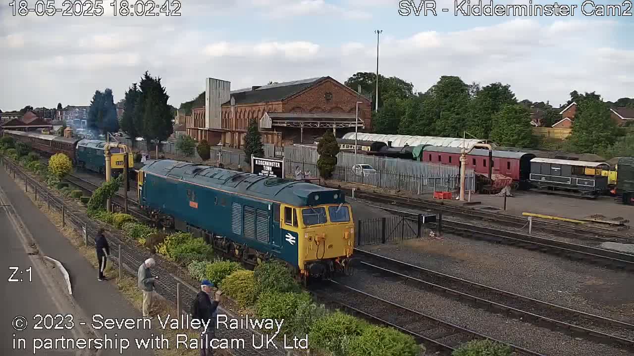 A teal and yellow diesel locomotive leads a train past a railway station on an overcast day.
