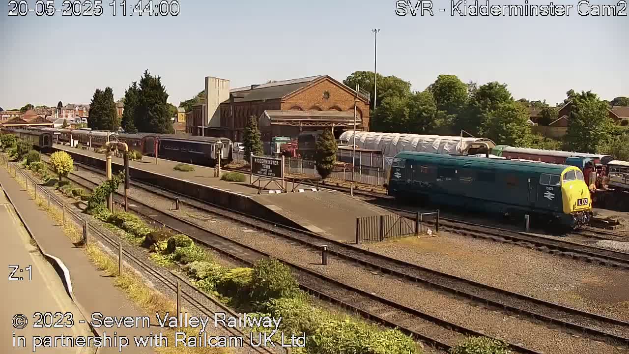 A sunny day at a train station shows several passenger cars and a teal diesel locomotive parked on train tracks, with additional rail cars visible in the background.