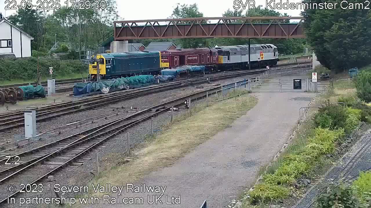 A train of several diesel locomotives and carriages sits on a railway track under a metal bridge on a partly sunny day.