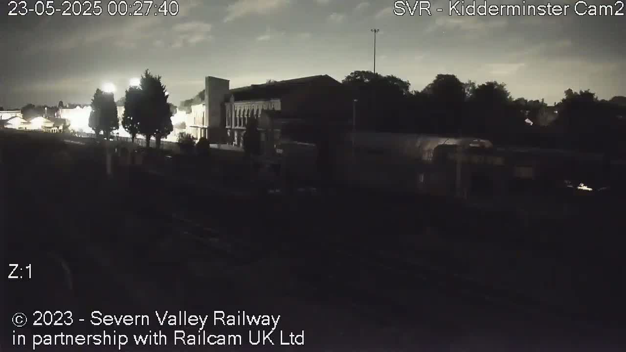 A dark, nighttime scene shows a train partially obscured by shadows near a building under a cloudy sky.