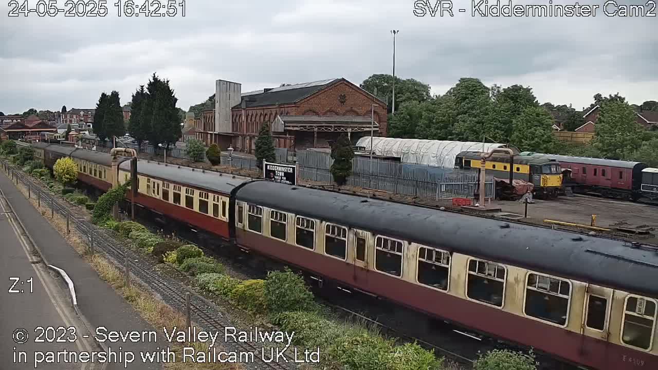 A long passenger train moves through a station on an overcast day, passing other railcars and a brick building.