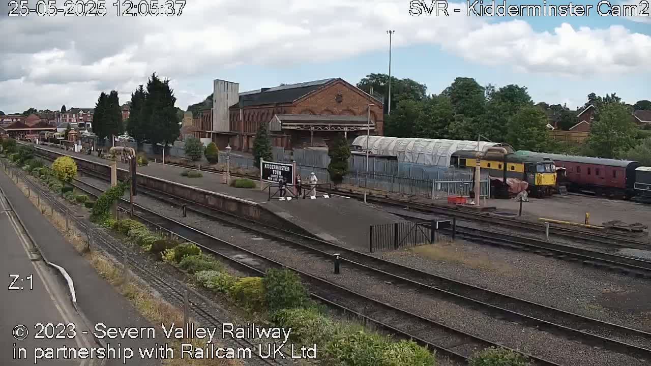 A railway station with several train cars and a brick building is visible under a partly cloudy sky.