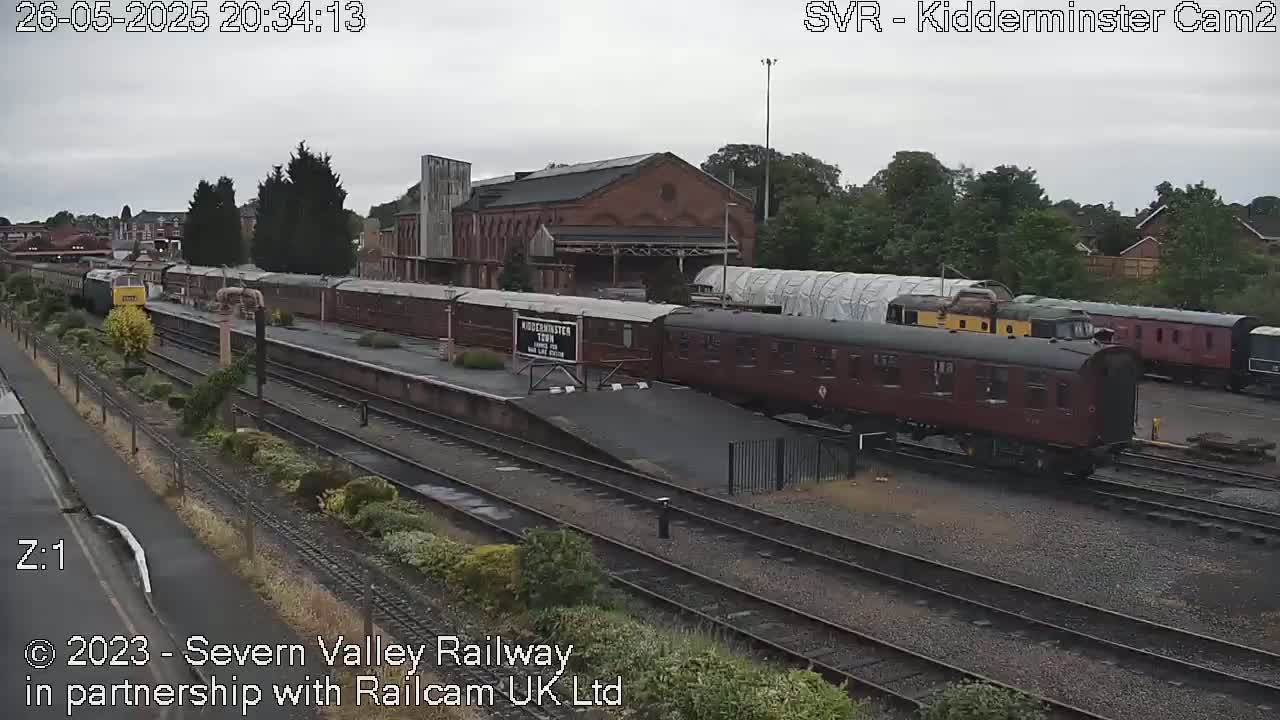 Under an overcast sky, a train sits at a station next to several railway cars and a large brick building.