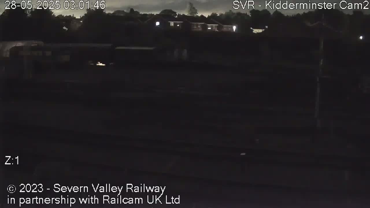A dark nighttime scene shows a train in a dimly lit rail yard under an overcast sky.