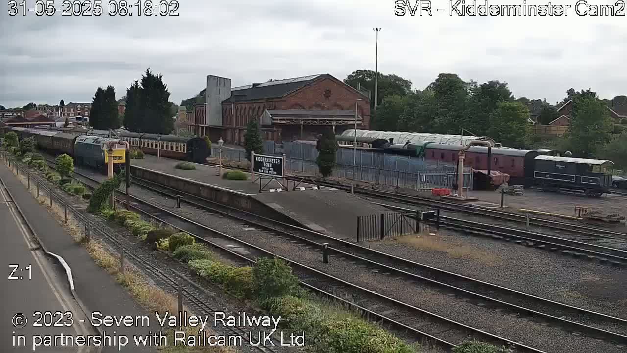 A train station with several passenger cars and locomotives is visible on an overcast day.