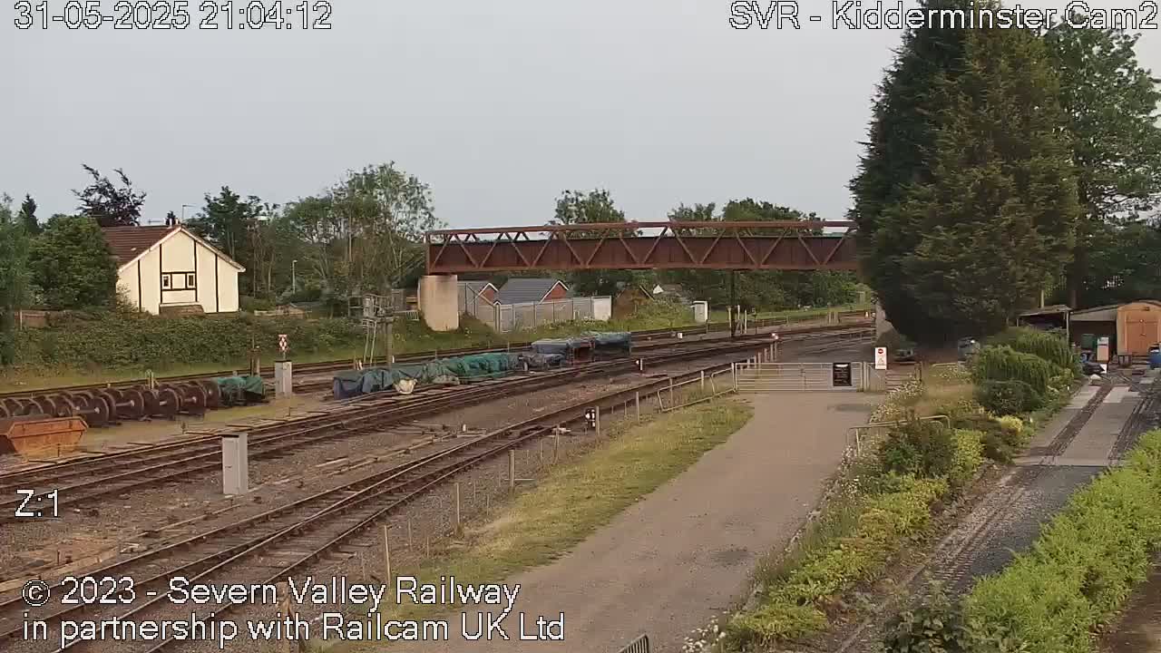 A railway yard with multiple train tracks, a metal footbridge, and several railway cars under a cloudy sky.