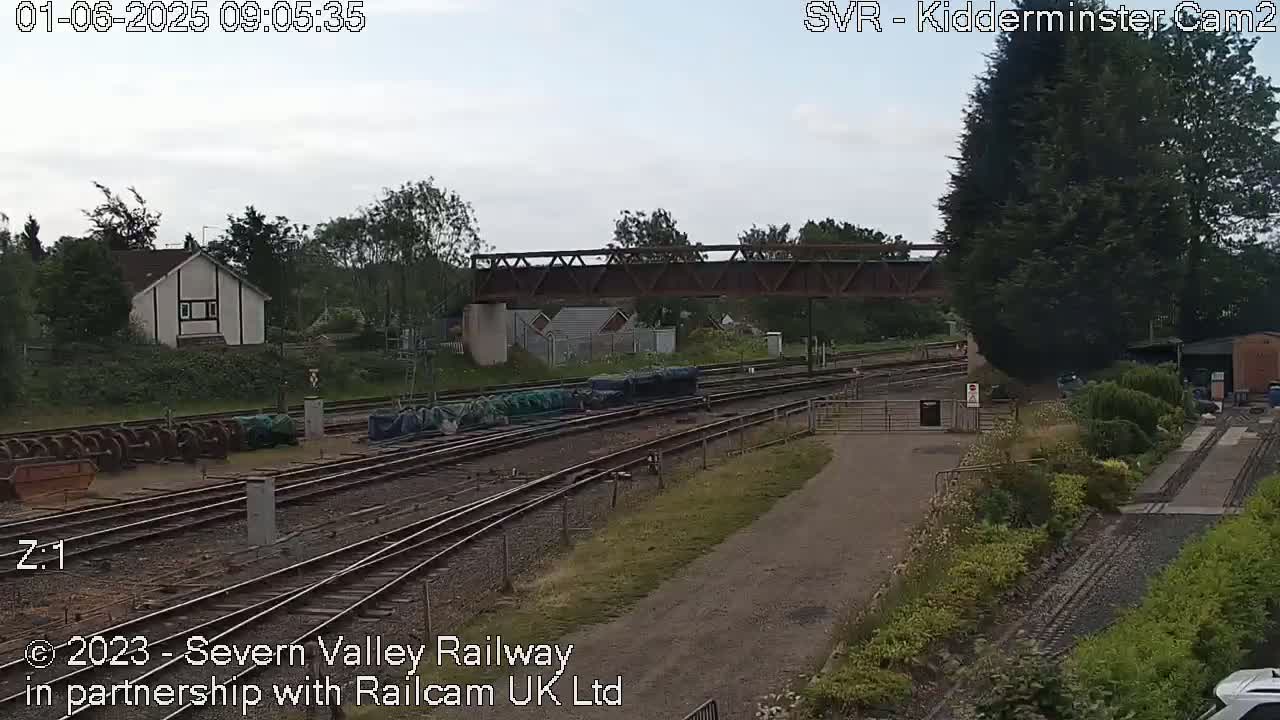A railway yard with multiple tracks, a metal bridge, and several buildings is visible under an overcast sky.