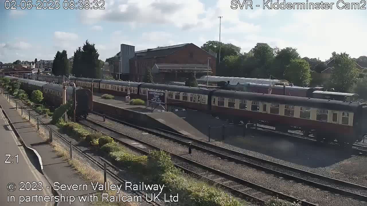 A partly cloudy day at a train station with several passenger cars and a locomotive on the tracks.