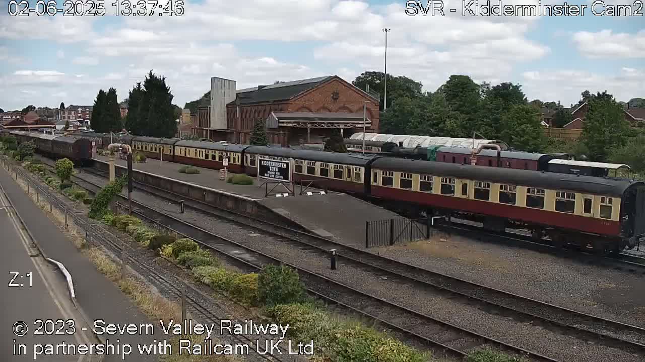 A long train of maroon and cream passenger cars sits at a station under partly cloudy skies.
