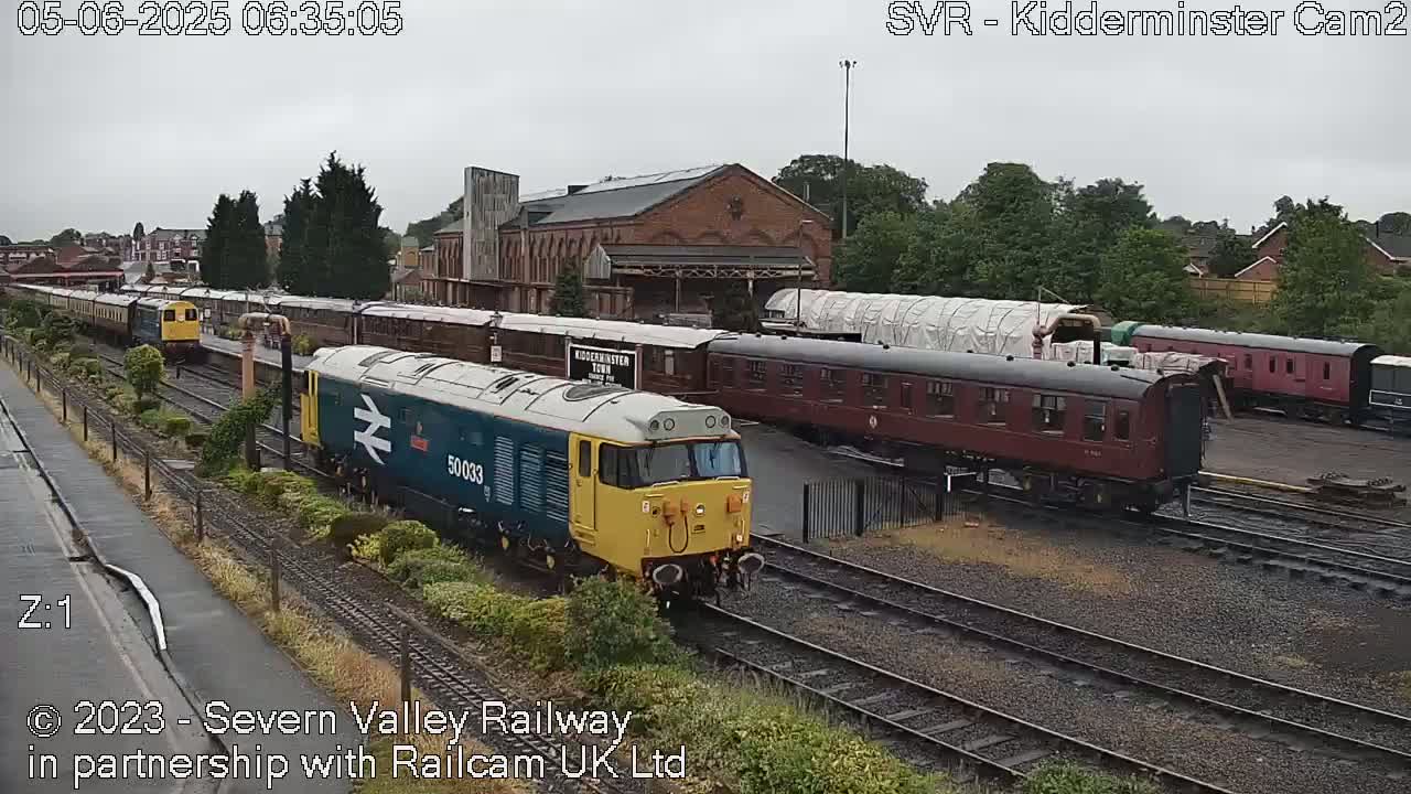 A blue and yellow diesel locomotive is moving past several stationary passenger cars on a railway line under an overcast sky.