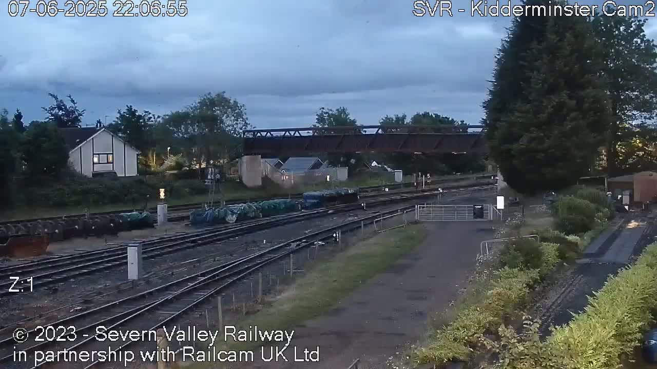 A railway yard under a cloudy evening sky, with several railway tracks, some covered rail cars, a bridge, and several buildings in the background.