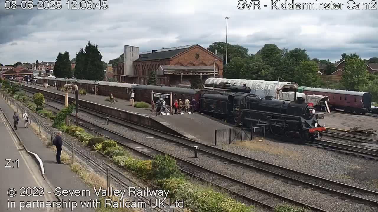 A black steam locomotive is parked at a train station on an overcast day, with several railway cars and people standing nearby.
