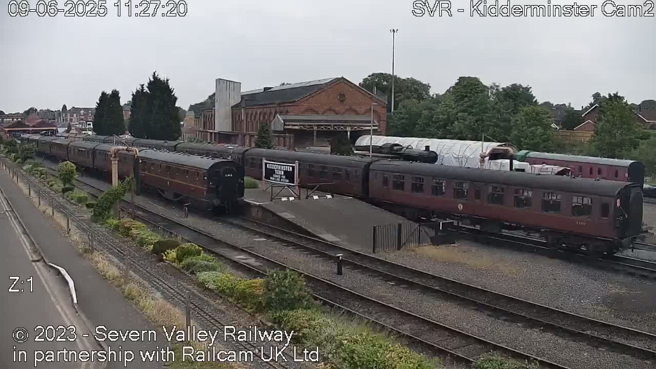 A train of maroon passenger cars sits in a railway station on an overcast day.