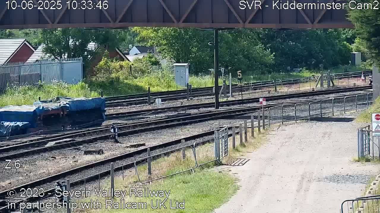 A sunny view of a railway yard with multiple tracks, a covered piece of railway equipment, and a dirt path leading away from the tracks.
