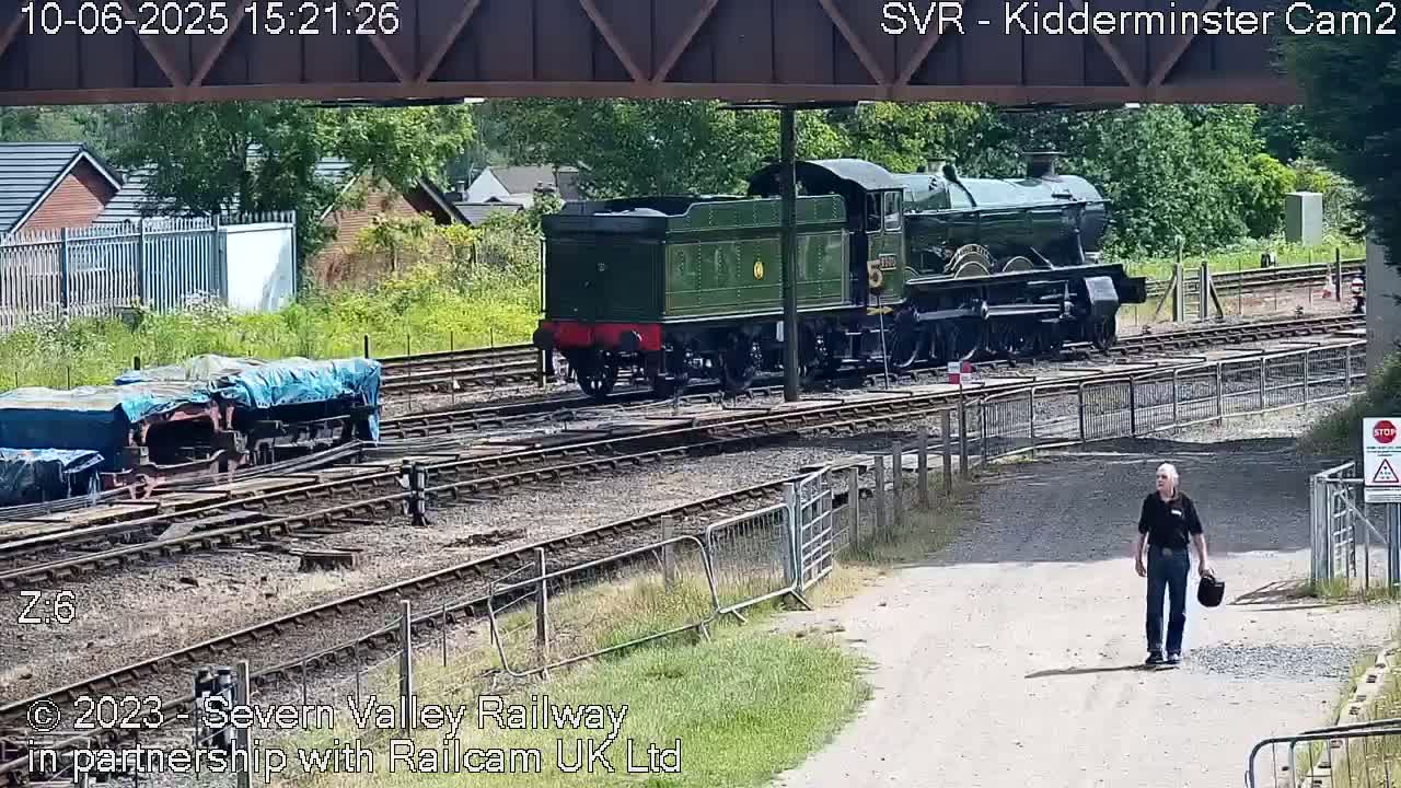 A green steam train is parked on railway tracks next to a covered railcar, with a man walking on a nearby path on a sunny day.